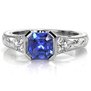 Kimberly's Dream is a lavish antique inspired ring displaying a rich asscher-cut blue sapphire in a half-bezel setting. This design features vintage inspired details including hand engraving, milgrain, and filigree. The cluster of three diamonds on either side of the center adds brilliance to its white gold mounting.