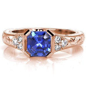 Kimberly's Dream is a lavish antique inspired ring displaying a rich asscher-cut blue sapphire in a half-bezel setting. This design features vintage inspired details including hand engraving, milgrain, and filigree. The cluster of three diamonds on either side of the center adds brilliance to its white gold mounting.