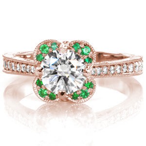 Vivid green emeralds outline four crescent shaped arches, creating a unique halo to showcase the center diamond. Micro pavé stones graduate as the band tapers. Hand engraved waves and filigree curls adorn the profile. The lotus style petal detail is embellished with diamonds for an antique look.