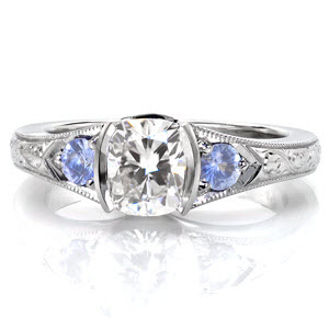 The pale blue sapphire sides stones of Design 3233, add a touch of serenity to this beautiful engagement ring design. The gently flared band turns into a half bezel setting as it reaches the cushion cut center diamond. The easy flowing curves of the band are elegantly adorned with hand engraving in a scroll pattern. 