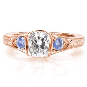Rose gold engagement ring in Oakland with cushion cut center stone, blue sapphire side stones and scroll hand engraving.