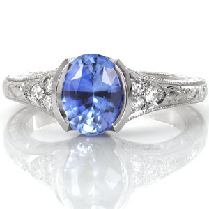 Design 3236 is shown with a 1.80 carat, oval cut, pale blue sapphire in a half bezel setting. The elongation of the center stone is accentuated by the flare of the band. This antique inspired engagement ring design is adorned with graceful hand formed filigree curls and elegant hand engraving. 