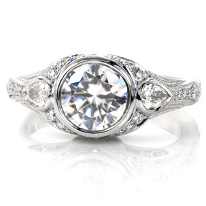 Crafted in Platinum, the Sylvia design has a full bezel center diamond with two pear cuts on each side. As the full flaired band tapers down, fine details of milgrain and round diamonds graduating in size adorn the band. Hand formed filigree curls are tucked under two grand swirls creating a great profile look.