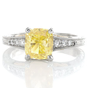 The vivid 1.70 carat fancy yellow cushion cut sapphire takes center stage in this vintage inspired ring. Yellow gold filigree curls complement the warm hue of the center stone. Ornate patterns are engraved on three faces of the ring for an antique finish. The edges are detailed with milgrain texture. 