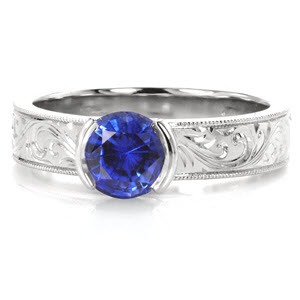 Design 3276 displays a beautiful 1.00 carat blue sapphire set within a half bezel setting. Creating a distinctive profile, the half bezel basket is joined with a wider band of 14k white gold.  Bright cut hand engraved scrolls catch just the right amount of light, hand applied milgrain edging frames the band design.