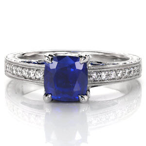 This elegant antique inspired design features a 1.00 carat cushion cut blue sapphire in four prongs. Hand engraving, diamonds and milgrain detail the sides of the ring for a vintage appeal. Marquise shaped petals are embellished with diamonds and open pockets are adorned with blue sapphires for a truly antique feel.