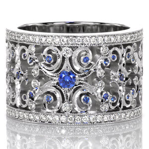 Sapphire Caledonia sets the stage for a regal look. Intricate openwork scrolls are accented with round cut diamonds and beautiful natural blue sapphires. Each scroll is finished with hand applied milgrain detail. The wide band is framed with outside rails of bead set round cut diamonds.