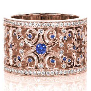 Sapphire Caledonia sets the stage for a regal look. Intricate openwork scrolls are accented with round cut diamonds and beautiful natural blue sapphires. Each scroll is finished with hand applied milgrain detail. The wide band is framed with outside rails of bead set round cut diamonds.