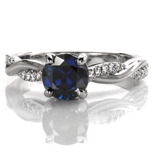The stunning 1.00 carat blue sapphire enhances the cool tone of this white gold ring. A row of round brilliant diamonds are interwoven between a high polish band, for dimension and contrast. Accent diamonds adorn the basket setting for brilliance. The profile features open pockets for a stylish side view.   