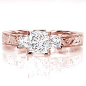 A classic three stone arrangement meets antique inspiration, for a truly one-of-a-kind look. The milgrain border creates a textured frame around the intricate scroll hand engraving. Chevron prongs are fashioned on the ends of the princess cuts for security and design appeal.     