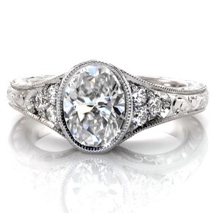 Design 3383 is a custom created engagement ring with antique inspirations. The oval center diamond is captured with a bezel setting that flows into the flared band. Delicate, hand formed filigree curls fill the pockets on the sides of the piece, while intricately carved hand engraving adorns the rest of the design.