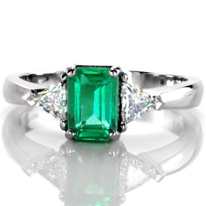 Design 3384 is a beautiful statement piece with a 1.20 carat emerald. The trillion side diamonds compliment the elongated, emerald cut shape of the center stone, while the band tapers in the meet the points of the diamonds. The vibrant, lush green stands out against the cool hues of the metal and diamonds. 