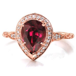 This stunning halo engagement ring is shown in 14 karat rose gold, featuring a 2.30 carat pear cut ruby center stone. The basket under the micro pavé halo features round diamond drapes and a bezel set marquise surprise diamond. The scalloped band is detailed with relief style hand engraving in a scroll pattern.