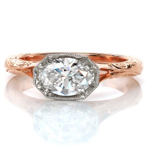 This stunning custom two-toned engagement ring design features a unique, horizontal oval center stone setting. The sides of the faux-bezel center setting are adorned with diamonds to form a horizontal halo. The rose gold split shank band is detailed with hand engraving and filigree curls for a vintage touch.
