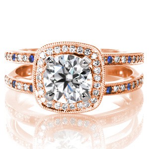 Custom Design 3469 offers a classic outline with the addition of dramatic blue sapphire side stones. A 1.00 carat round brilliant diamond is held at the center surrounded by a bead set diamond halo. Blue sapphire are staggered across the split shank diamond band. Milgrain edging adds the finishing touch to this ring.