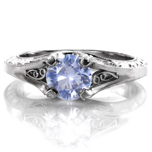 Design 3475 is a stunning vintage inspired custom engagement ring that features an unusual and extraordinary 0.80 carat round light blue sapphire. A four prong setting holds the cornflower blue sapphire with elegant hand formed filigree curls and delicate hand engraving surrounding the center stone.  