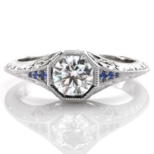 Design 3487 is a stunning, vintage inspired engagement ring featuring a round brilliant center diamond. The knife edge band flares as it approaches the octagonal center setting to reveal bead-set blue sapphires. Hand-formed filigree curls, and elegant hand engraved half-wheat patterns adorn the sides of the band.