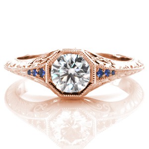 Design 3487 is a stunning, vintage inspired engagement ring featuring a round brilliant center diamond. The knife edge band flares as it approaches the octagonal center setting to reveal bead-set blue sapphires. Hand-formed filigree curls, and elegant hand engraved half-wheat patterns adorn the sides of the band.