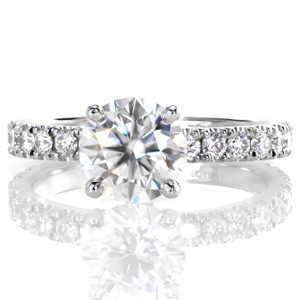 Round brilliant diamonds shine from every angle of this gorgeous design. Hand formed u-cuts minimize the metal holding the stones while securely fashioning the diamonds to provide ultimate sparkle. The four prong setting features a 1.30 carat round center and an apron of diamonds between each prong.