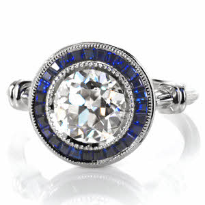 Custom Design 3518 is truly a show-stopper with its dazzling 2.00 carat round center diamond and deep blue sapphire tapering banquette framing halo. This vintage inspired custom ring features milgrain edging, wrapped shoulders, and a pierced under basket. 