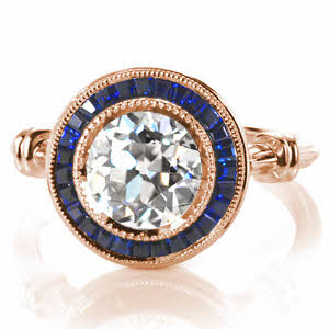 Custom Design 3518 is truly a show-stopper with its dazzling 2.00 carat round center diamond and deep blue sapphire tapering banquette framing halo. This vintage inspired custom ring features milgrain edging, wrapped shoulders, and a pierced under basket. 