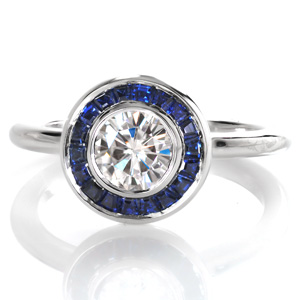 Design 3527 is a captivating custom engagement ring with clean lines and gemstone halo. A glittering 0.70 carat round diamond is held in a full bezel setting and surrounded by a custom halo comprised of perfectly placed tapered baguette blue sapphires. This impressive gemstone halo sits atop a classic rounded band.