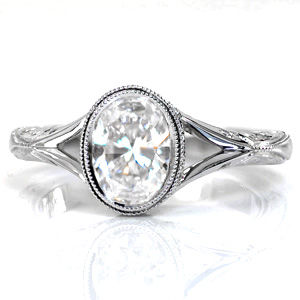 This artistic custom design features a bezel set oval center stone bordered with a double row of milgrain. The delicate split shank tapers into a hand engraved band. A decorative side halo frames the setting, with flush-set diamonds on the split of the shank as well. Hand wrought filigree curls complete this ring.