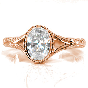 This artistic custom design features a bezel set oval center stone bordered with a double row of milgrain. The delicate split shank tapers into a hand engraved band. A decorative side halo frames the setting, with flush-set diamonds on the split of the shank as well. Hand wrought filigree curls complete this ring.