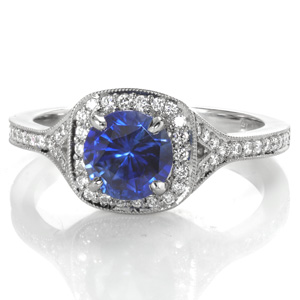 A lush, blue sapphire adds a regal air to this modern halo engagement ring design. A narrow diamond band flares and separates near the top to seamlessly flow into the diamond halo. A beaded texture called milgrain has been hand applied to the edges for a sophisticated look.