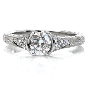 This organically inspired engagement ring features a low, half-bezel setting with a flared band. The spectacular relief engraving on the top and sides of the piece takes form as delicate flowers and flowing, curling vines. Hand wrought filigree curls fill the pockets to either side of the center stone.