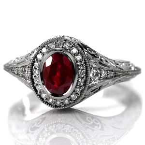 Design 3623 is shown featuring a bezel set 1.20 carat oval cut ruby center stone. A micro pavé halo frames the gem and blends seamlessly into the flared knife edge band. The sides of the band are elegantly detailed with relief style hand engraving and pockets of hand wrought filigree curls.