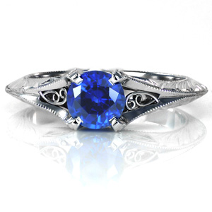 Elegant antique inspired details come together to create a stunning solitaire engagement ring featuring a lush blue sapphire. The flared knife edge band is ornately detailed with hand engraved patterns and hand wrought filigree curls. The band flows up to form the center prongs with milgrain textured edges. 