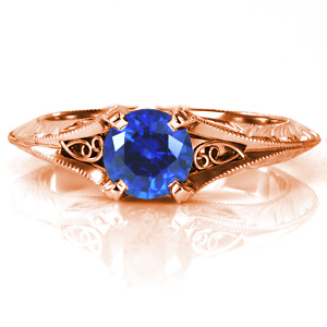 Elegant antique inspired details come together to create a stunning solitaire engagement ring featuring a lush blue sapphire. The flared knife edge band is ornately detailed with hand engraved patterns and hand wrought filigree curls. The band flows up to form the center prongs with milgrain textured edges. 