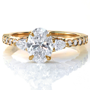 A 1.0 carat oval diamond is flanked by two pear shaped diamonds in Design 3707. The unique band features bezel set round diamonds and a high polish finish. A glamorous five petal diamond detail adds extra sparkle and flair under the custom center setting. 