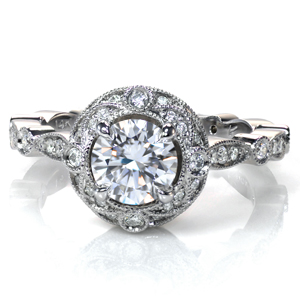 A one carat diamond is surrounded by a unique, scalloped double halo featuring beadset and bezel set diamonds. The band also features a scalloped design and is accented by more diamonds. Milgrain detail is applied by hand to the halo and the band, and filigree curls are placed under the basket for more vintage flair. 