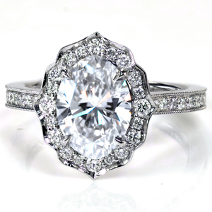 Shown with a 1.5 carat oval cut diamond, this majestic engagement ring features a scalloped halo set with mixed sizes of round brilliant cut diamonds. The cathedral style band is adorned with more sparkling round diamonds and accented with hand applied milgrain detail. 