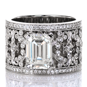 Design 3755 is a stunning example of sophisticated artistry. A 2 carat emerald cut diamond is set atop the intricately detailed wide band. Broad filigree flourishes curl and hold bezel set round diamonds. Each turn of metal has hand applied milgrain detail for a vintage look. Rows of bead set diamonds frame the band. 