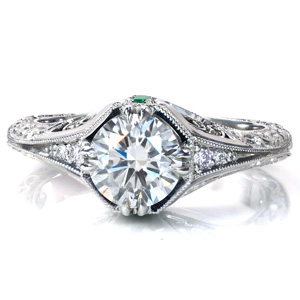 A 1.20 carat diamond is framed by 4 sets of double prongs and a unique under halo in Design 3765. The flared band features graduated diamonds leading to a knife edge, intricate relief style engraving, and hand applied milgrain detail. This vintage inspired beauty is finished with filigree curls and a surprise emerald.