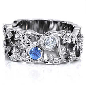 This unique wide band features a floral motif, incorporating swirling vines and leaves. The flowers are woven throughout the ring, and the vines wrap around the sapphire and diamond in bezel settings. A double row of milgrain detail is applied to the vines, adding to the vintage feel of this ring. 