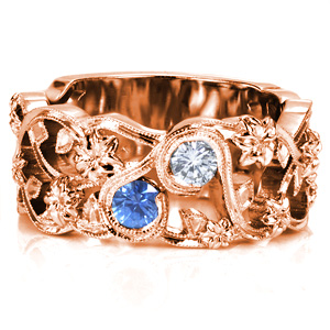 This unique wide band features a floral motif, incorporating swirling vines and leaves. The flowers are woven throughout the ring, and the vines wrap around the sapphire and diamond in bezel settings. A double row of milgrain detail is applied to the vines, adding to the vintage feel of this ring. 