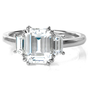 A stunning 2 carat emerald cut diamond is nestled in a low basket setting, held by four sets of double claw prong. Unique trapezoid diamonds grace each side of the center stone, a lovely update to the classic three stone design. A thin, high polished band completes this elegant engagement ring. 