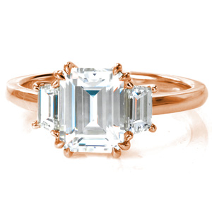 A stunning 2 carat emerald cut diamond is nestled in a low basket setting, held by four sets of double claw prongs. Unique trapezoid diamonds grace each side of the center stone, a lovely update to the classic three stone design. A thin, high polished band completes this elegant engagement ring. 