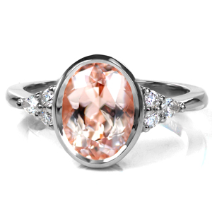 A secure bezel setting protects the delicate, peachy pink morganite center stone. The open gallery under the center setting is decorated with hand formed filigree curls. Clusters of round brilliant and pear shaped diamonds are set atop the slightly raised shoulders, meeting the high polished band at a slight taper. 