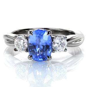 A mesmerizing oval shaped blue sapphire is flanked by two oval diamonds in this elegant design. Each stone is hand set in a classic four prong style, and a delicate, draped basket adds a lovely touch to the profile. The high polished band appears intertwined for added appeal. 