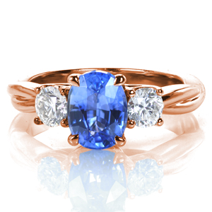 A mesmerizing oval shaped blue sapphire is flanked by two oval diamonds in this elegant design. Each stone is hand set in a classic four prong style, and a delicate, draped basket adds a lovely touch to the profile. The high polished band appears intertwined for added appeal. 