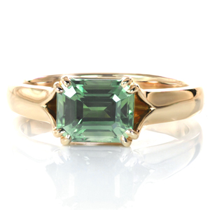 A rich green emerald cut sapphire is the star of this design. The east-west setting of the center stone adds a modern flair to a classic solitaire setting. The high polished band flares and opens toward the center stone, allowing light in from all sides. Four sets of small double claw prongs complete the look. 