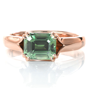 A rich green emerald cut sapphire is the star of this design. The east-west setting of the center stone adds a modern flair to a classic solitaire setting. The high polished band flares and opens toward the center stone, allowing light in from all sides. Four sets of small double claw prongs complete the look. 