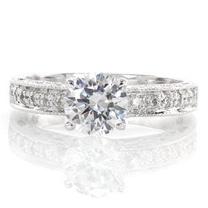 The Adorable design is simply charming! A .80 carat round center diamond is accented by bead-set side stones on the top of the band. The sides feature bezel set diamonds and cut outs, including a heart shape on the side of the center stone. All the edges are detailed in milgrain to add texture. 